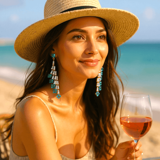 Woman on a beach holding a glass of pink wine, wearing a straw hat and large earrings.