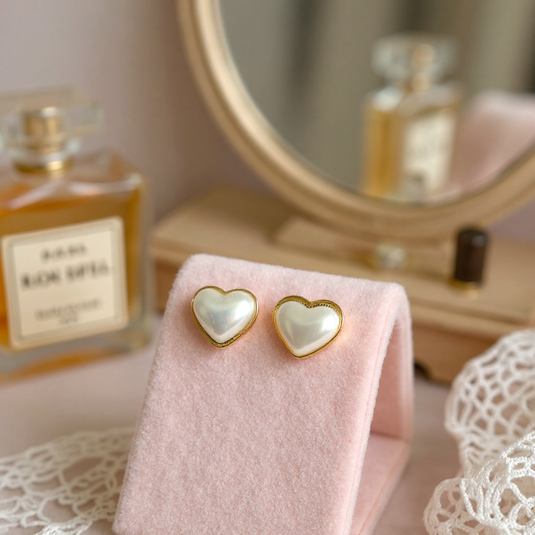 Heart-shaped pearl earrings on a pink cloth with a perfume bottle in the background